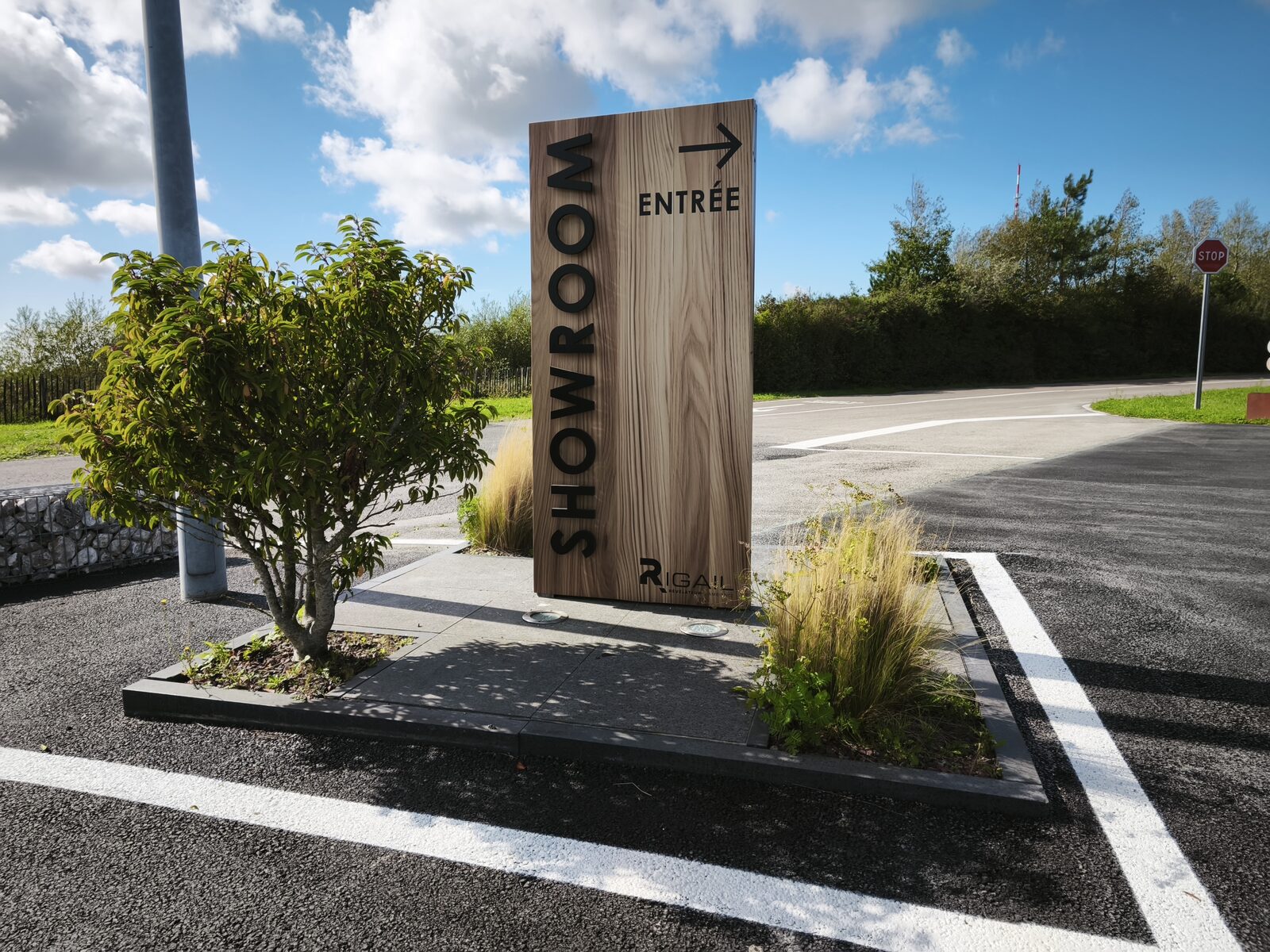 Totem « Showroom — Entrée » habillage effet bois, posé dans un massif paysager du parking du showroom Rigail à Saint-Martin-Boulogne